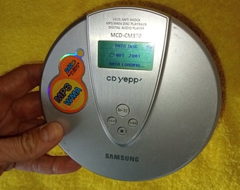 May include: A silver Samsung MCD-CM370 CD player with a black and white LCD screen displaying "DATA DISC 801 2001 DATA LOADING..." and the text "CD Yepp" below. The player has a silver face with buttons for play, pause, stop, forward, and rewind. The player has a sticker on the side that says "MP3 WMA 240sec Anti-Shock".