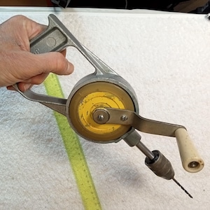 May include: A hand-crank drill with a yellow and grey handle and a white plastic handle. The drill bit is visible.