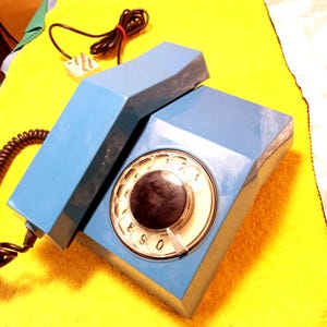 May include: A vintage blue rotary dial telephone with a black dial and a coiled cord. The phone has a rectangular base and a handset. The phone is on a yellow surface.