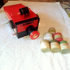 May include: A vintage red and black Kwant slide projector with a black lens and a series of small jars. The projector has a rectangular shape with a top handle and a power cord. The jars are various colors and have labels.