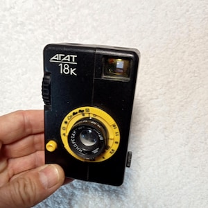 Op de afbeelding: Een zwarte vintage AGAT 18K camera met een gele lensring en wijzerplaat. De camera heeft een rechthoekige vorm met een lens, flitser en opwindknop. De tekst "AGAT" en "18K" staan op de voorkant van de camera.