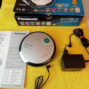 May include: A silver and black Panasonic SL-CT490 portable CD player with a green button and a headphone jack. The player is on a yellow surface with a black power adapter and a black headphone cable.