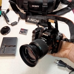 Pode incluir: Câmera DSLR Sony Alpha preta com lente, alça e bolsa de transporte. Inclui carregador, tampa da lente e outros acessórios. O logotipo da Sony é visível na câmera e na lente.