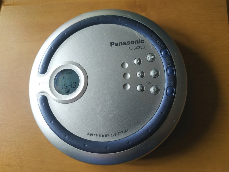 Puede incluir: Un reproductor de CD port&aacute;til plateado y azul con una pantalla digital que muestra "5 16:30". El reproductor tiene botones para el volumen, el salto y otras funciones. El texto "Panasonic SL-SX320" y "Anti-Skip System" est&aacute; impreso en el reproductor.