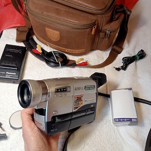 Peut inclure: Caméscope Panasonic RZ10 argenté et noir avec sac de transport en cuir marron, chargeur et accessoires. Le caméscope a un zoom numérique 250x. Une mini-cassette DV est également incluse.