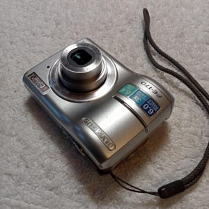 May include: Silver Olympus FE-170 digital camera with a black lens and a dark gray wrist strap. The camera has a 6.0-megapixel resolution and 3x zoom. The camera has the Olympus logo on the front.