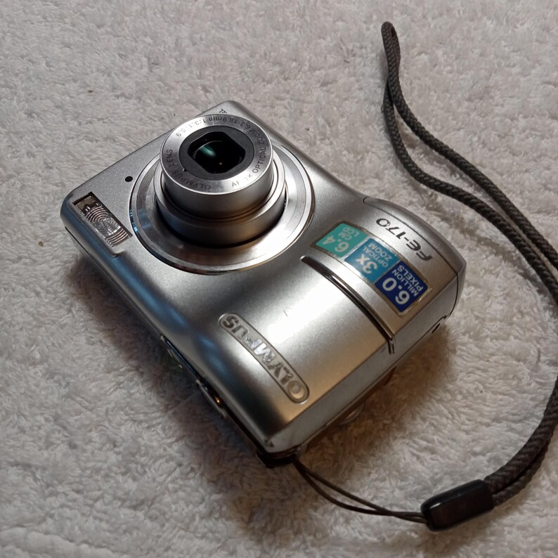 May include: Silver Olympus FE-170 digital camera with a black lens and a dark gray wrist strap. The camera has a 6.0-megapixel resolution and 3x zoom. The camera has the Olympus logo on the front.