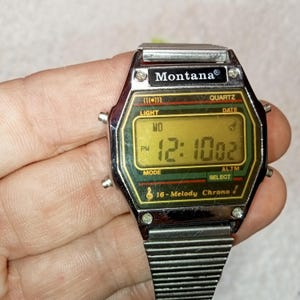 May include: A silver Montana digital watch with a rectangular face showing the time 12:10:02 PM. The watch has a black and green border with the text "Montana" at the top. The watch strap is silver and has a ribbed design.