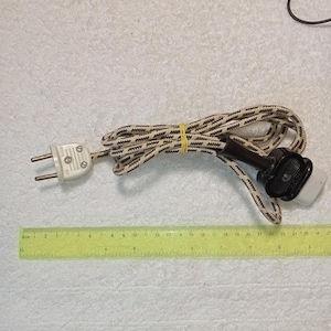 Può includere: Cavo elettrico con motivo a zig-zag bianco e marrone. Ha una spina bianca con due pin e una presa nera e bianca. Un elastico giallo tiene insieme il cavo. Misure in cm.