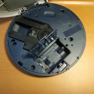 Puede incluir: Primer plano de un reproductor de CD azul con la bandeja del disco abierta, revelando el mecanismo interno y una ranura para un disco. El reproductor est&aacute; sobre una superficie de madera.