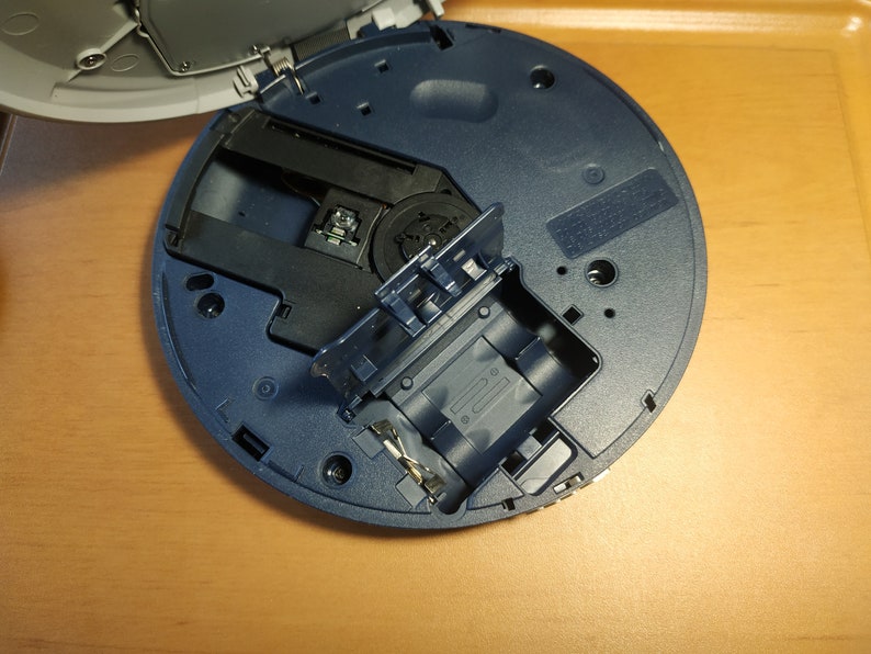 Puede incluir: Primer plano de un reproductor de CD azul con la bandeja del disco abierta, revelando el mecanismo interno y una ranura para un disco. El reproductor est&aacute; sobre una superficie de madera.