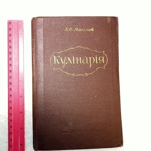 May include: A vintage brown hardcover cookbook with gold lettering and the title "(Kулiнарiя)". The author's name, "Л.О. Маслов", is above the title. A pink ruler is on the left side of the book, showing measurements in inches.