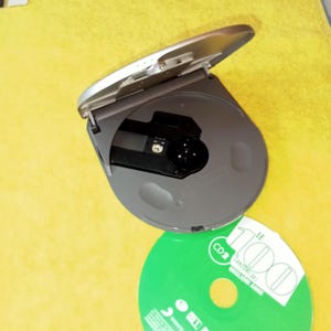 May include: A silver CD player with the lid open, revealing the disc tray. A green CD is next to the player, with the text "LE 1000" and "CD 2" printed on it.