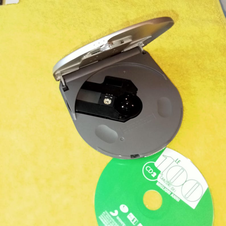 May include: A silver CD player with the lid open, revealing the disc tray. A green CD is next to the player, with the text "LE 1000" and "CD 2" printed on it.