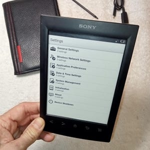 Puede incluir: Un lector electrónico Sony negro que muestra el menú de configuración, con opciones para la configuración general, la configuración de red inalámbrica y las preferencias de la aplicación. También se ve una funda de cuero negra y un lápiz óptico.