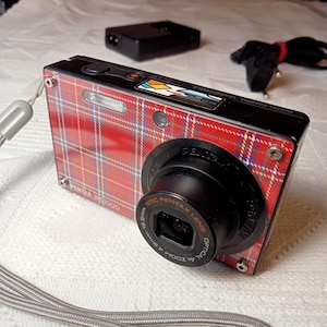 Op de afbeelding: Een rood-zwarte digitale camera met een geruit ontwerp. De camera heeft een 4x optische zoomlens en een SMC Pentax lens. De camera is gelabeld met "Digitale camera" en "A Megapixels".