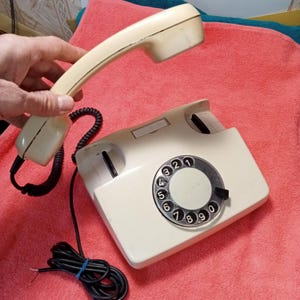 May include: An off-white vintage rotary dial telephone with a matching handset. The phone has a circular dial with black numbers and a black cord. The handset is held up, showing the receiver and mouthpiece. The phone is on a pink surface.