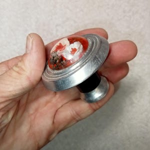 May include: A decorative bottle stopper with a silver-coloured metal base and a clear, rounded top. The top features a red and white floral design with a black stem. The stopper is designed to fit into a bottle.