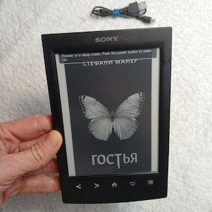Puede incluir: Un lector electrónico Sony negro con una pantalla rectangular que muestra un gráfico de mariposa y el texto "Reader is in sleep mode. Push the power button to wake up." El dispositivo tiene botones de navegación y un cable USB negro.