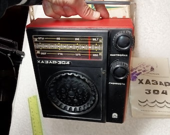 May include: Vintage black and red radio with tuning dials and a speaker grill. The radio has the text "ХАЗАР-304" on the front. A yellow ruler is visible in the image. A manual is also present.
