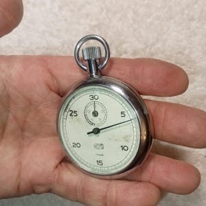 Op de afbeelding: Een vintage zilveren stopwatch met een witte wijzerplaat en zwarte cijfers. Het horloge heeft een kleine subwijzerplaat en een grote secondewijzer. De tekst "UMF Ruhla" is zichtbaar op de wijzerplaat. De stopwatch wordt in een hand gehouden.