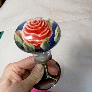 May include: A decorative bottle stopper with a red rose design encased in a glass orb. The orb has a blue background with green leaves. The stopper has a silver-coloured stem and base.