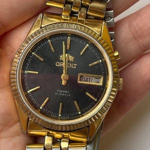 Può includere: Orologio Orient vintage con cassa e cinturino dorati. Il quadrante dell'orologio è nero con lancette e indicatori dorati. L'orologio mostra il giorno e la data. Il cinturino è una combinazione di maglie dorate e argentate.