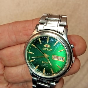 May include: A silver Orient wristwatch with a green face. The watch shows the day and date, with "SUN 11" visible. The watch has a stainless steel strap and a crystal face. The watch is held in a hand.
