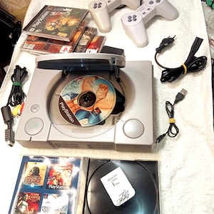 Peut inclure: Une console PlayStation argentée avec un disque Disney's Hercules. Comprend deux manettes blanches, des disques de jeu et une carte mémoire. Des câbles et des cordons d'alimentation sont également visibles.