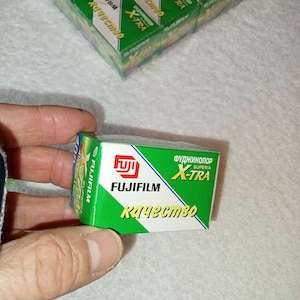 Op de afbeelding: Een groene en witte Fujifilm Superia X-TRA filmdoos. De doos heeft het Fujifilm-logo en de tekst "X-TRA" in het geel. De doos wordt in een hand gehouden.