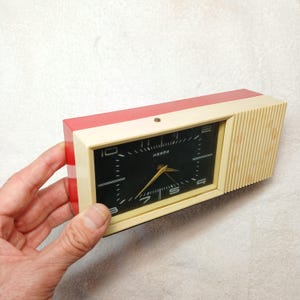 May include: A vintage rectangular alarm clock with a red top, cream-colored frame, and a black clock face. The clock face has white markings and gold hands. The clock is being held by a hand.