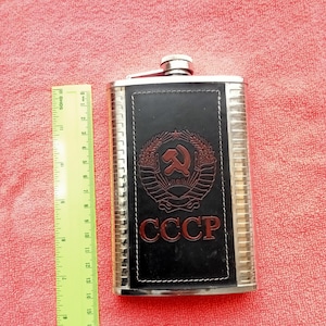 May include: A stainless steel flask with a black leather panel featuring a red emblem and the letters "CCCP". The flask is rectangular with a hinged cap and measures approximately 5.5 inches tall.