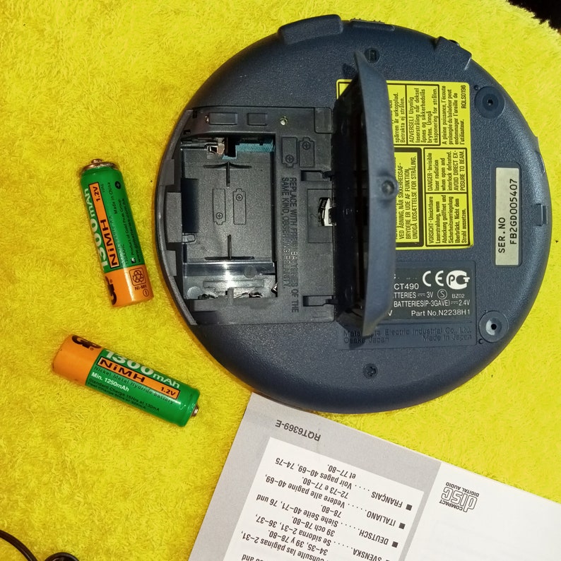 May include: A blue portable CD player with a battery compartment open, revealing two AA batteries. The player has a CE mark and a serial number FB2GD005407. Two green AA batteries are visible on a yellow surface.
