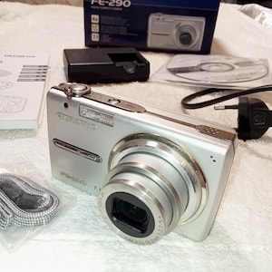 May include: Silver Olympus FE-290 digital compact camera with a lens, flash, and the Olympus logo. Accessories include a charger, a CD, and a camera strap. The camera box is visible in the background.