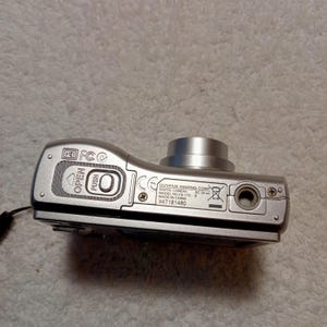 May include: Silver Olympus FE-170 digital camera. The camera has a lens, a flash, and several buttons. The bottom of the camera has the text "OLYMPUS IMAGING CORP DIGITAL CAMERA MODEL NO. FE-170 E MADE IN CHINA X47181460".