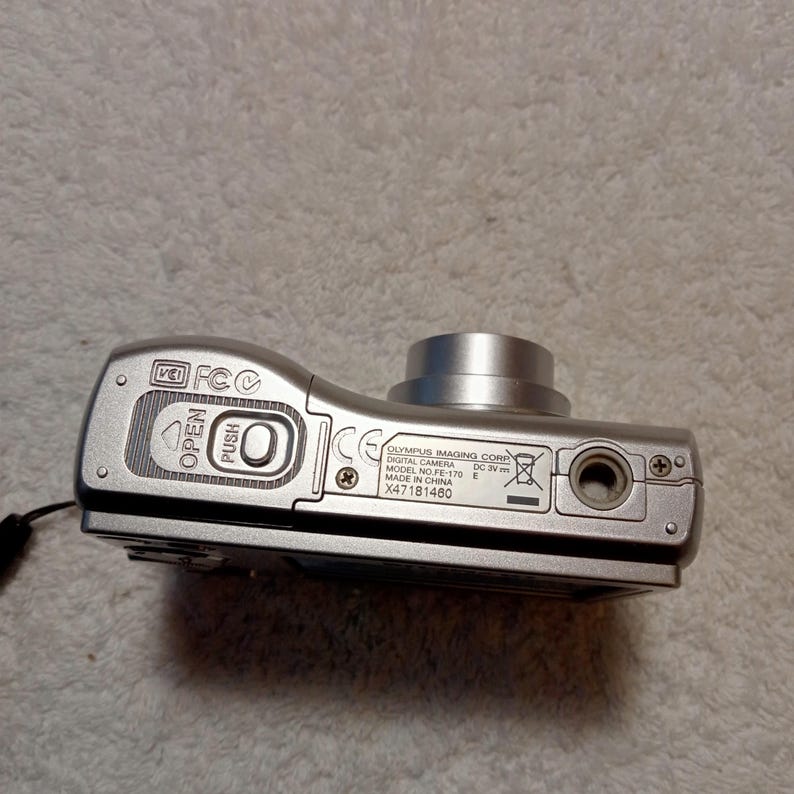 May include: Silver Olympus FE-170 digital camera. The camera has a lens, a flash, and several buttons. The bottom of the camera has the text "OLYMPUS IMAGING CORP DIGITAL CAMERA MODEL NO. FE-170 E MADE IN CHINA X47181460".