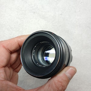 Peut inclure: Un objectif d'appareil photo noir avec des d&eacute;tails argent&eacute;s, tenu dans une main. L'objectif porte des inscriptions, dont "2/58" et "HELIOS-44-2". Le verre refl&egrave;te la lumi&egrave;re, et l'objectif est net. Accessoire de photographie.