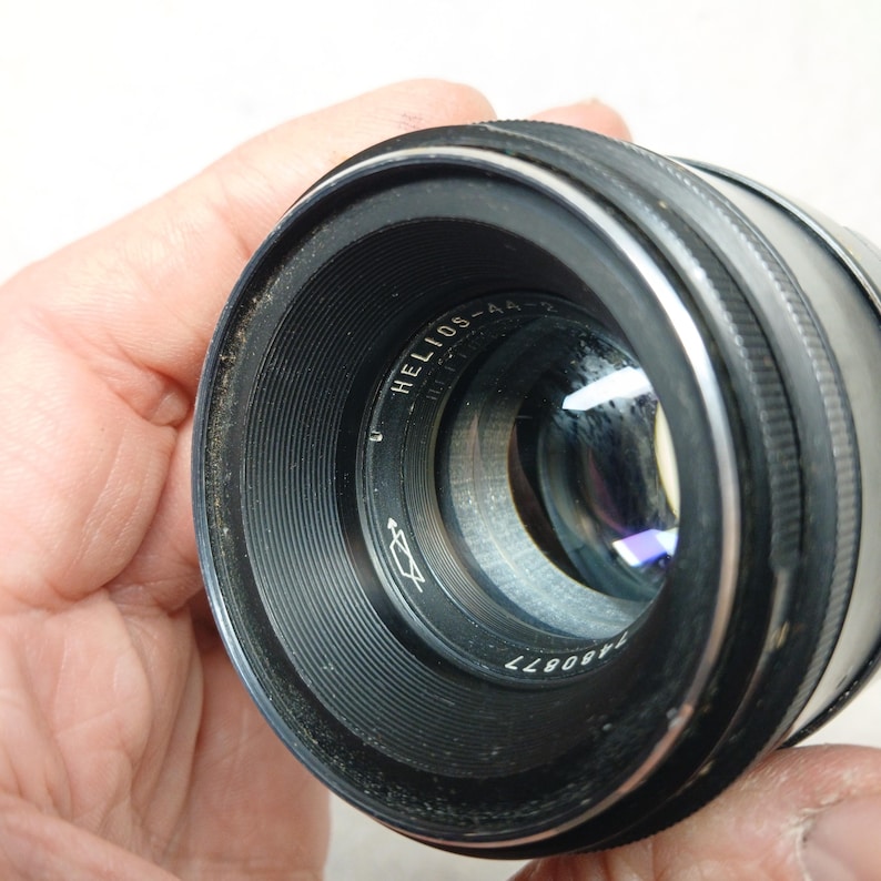 Peut inclure: Un objectif d'appareil photo noir Helios-44-2 avec une bague argent&eacute;e et une lentille en verre. L'objectif porte l'inscription "HELIOS-44-2" et un num&eacute;ro de s&eacute;rie. L'objectif est tenu dans une main.