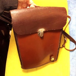 May include: A brown leather satchel with a flap closure and a shoulder strap. The bag has a rectangular shape with a rounded top and a silver-colored clasp. The leather appears worn, giving it a vintage look. The bag is held by a person.