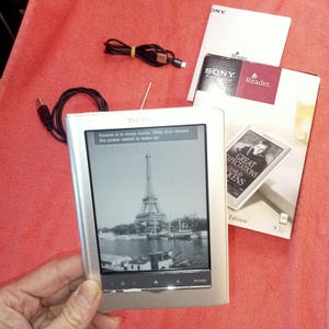 Puede incluir: Lector electrónico Sony plateado que muestra una imagen en blanco y negro de la Torre Eiffel. La pantalla dice "Reader is in sleep mode, slide and release the power switch to wake up." Incluye un cable USB y materiales impresos.