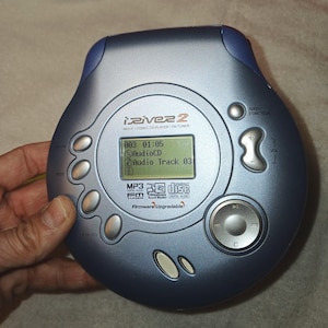 Può includere: Un lettore CD MP3 iRiver blu e argento con un piccolo schermo LCD che mostra le informazioni della traccia. Il lettore ha diversi pulsanti e una manopola di navigazione. Il testo "iRiver" e "MP3" sono visibili.