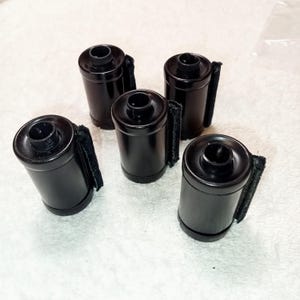 May include: Five black film canisters with a matte finish are arranged on a white surface. Each canister has a small, black fabric tab attached. The canisters are cylindrical with a slightly wider top.