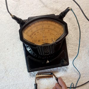 Puede incluir: Un multímetro analógico TR-4 negro vintage con un indicador circular y una aguja. El indicador tiene marcas y números. Los cables están conectados al multímetro y a una batería Panasonic. El dispositivo está sobre una superficie blanca.