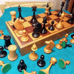 Op de afbeelding: Een houten schaakspel met zwarte en lichtgekleurde stukken op een schaakbord. De stukken zijn opgesteld voor een spel, met enkele stukken verspreid rond het bord. De set staat op een blauw oppervlak.