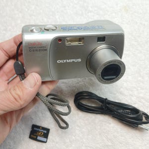 May include: A silver Olympus Camedia C-310 Zoom digital camera with a lens, flash, and wrist strap. The camera body features the text Olympus, Camedia, and C-310 Zoom. Included are a black USB cable and a small black Olympus xD-Picture Card.