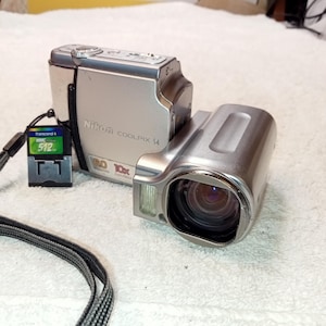 Puede incluir: Cámara digital plateada Nikon Coolpix 14 con objetivo zoom 10x. Incluye una tarjeta de memoria de 512 MB y una correa negra. La cámara tiene la inscripción "Nikon COOLPIX 14" en la parte frontal.