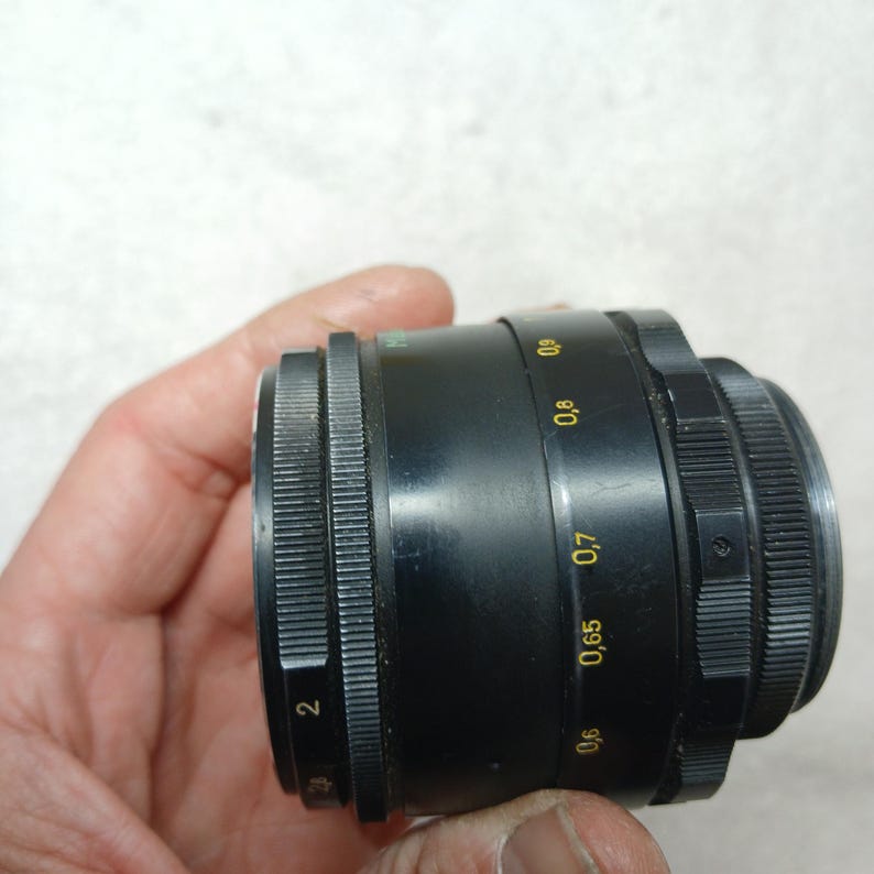 Peut inclure: Un objectif d'appareil photo noir avec une bague de mise au point stri&eacute;e et des marquages de distance en jaune et vert. L'objectif porte le marquage 2 sur la bague de mise au point. L'objectif est tenu dans la main d'une personne.