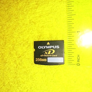 Puede incluir: Una tarjeta xD-Picture Olympus negra con texto blanco y un triángulo dorado. La tarjeta dice "OLYMPUS", "xD", "xD-Picture Card" y "256MB". Una regla amarilla está en el fondo.