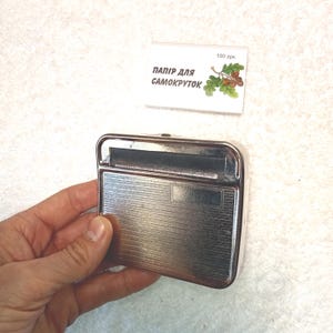 May include: A silver metal cigarette rolling machine with a black plastic roller. The machine has a textured surface and is in the shape of a rectangle. The text "ПАПІР ДЛЯ САМОКРУТОК" is visible on a white label in the background.
