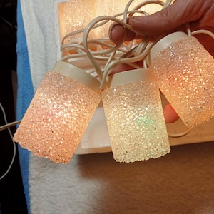 May include: A string of vintage Christmas lights with textured, cylindrical bulbs. The bulbs are a frosted, speckled plastic, emitting a warm, soft glow. The string is held by a hand, showcasing the off-white cords and bulb bases.